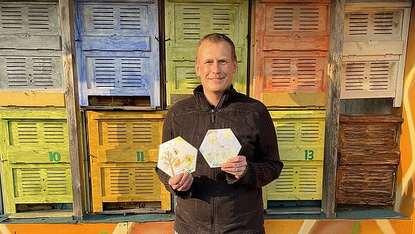 Imker Stefan Potraz steht vor den bunt bemalten Bienenstöcken und hält die beiden Bienenwaben in die Kamera, die aus der neu.sw Patenklasse stammen. 