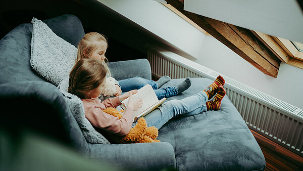 Zwei Mädchen sitzen auf einem Sofasessel, der an einer Heizung steht, und lesen ein Buch.