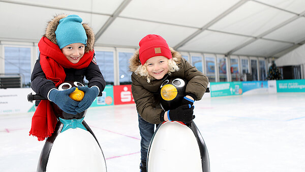 Zwei Kinderstützen sich auf Eislaufhilfen in Form von Pinguinen auf der Eislauffläche der neu.sw Eislaufhalle. 