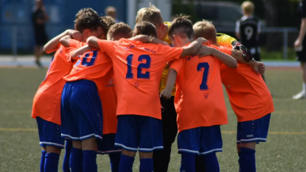 Fußball-Kinder stehen Arm in Arm im Kreis. Sie tragen orange und dunkelblau.