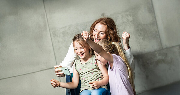 Frau und zwei kleine Mädchen jubeln beim Blick auf ein Smartphone. 