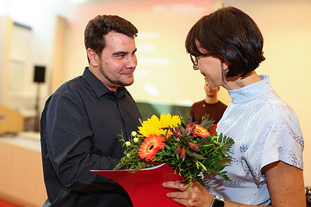 Frau überreicht einem Mann Blumen und Urkunde.