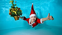 Ein kleiner Junge mit Weihnachtsmütze schwimmt unter Wasser mit ausgestreckten Armen, und einem Weihnachtsbaum in der rechten Hand. Er blickt in die Kamera und lächelt. 