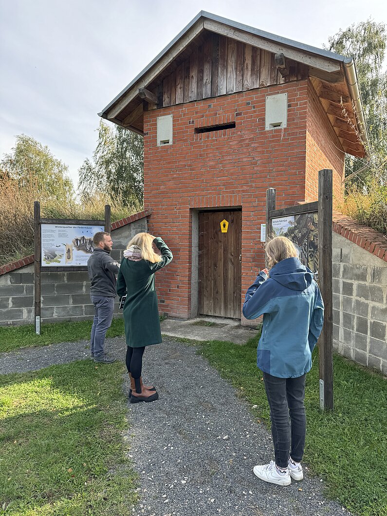 Drei Menschen stehen vor einem Backsteinhaus mit Einflugschlitzen für Fledermäuse. An der Tür ein Naturschutz-Schild mit schwarzer Eule auf glebem Grund.