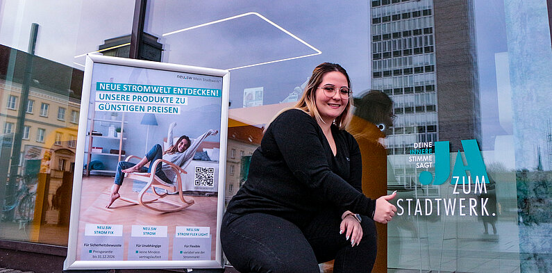 Junge Frau sitzt vor Schaufenster des neu.sw Kundenbüros und zeigt lächelnd auf die Beklebung "JA zum Stadtwerk." Im Schaufenster spiegelt sich der HKB-Turm als höchstes Gebäude am Marktplatz.