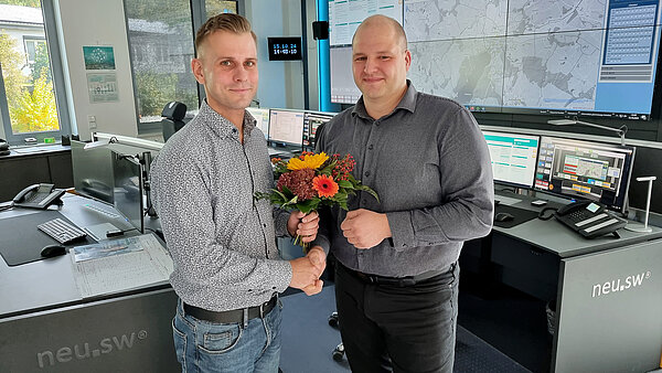 Betriebsleiter Alexander Pahlke (links) gratuliert Ingenieur Gordon Lange mit einem Blumenstrauß. Beide stehen in der Leitzentrale vor einer großen Bildschirmwand mit technischen Anzeigen..