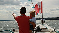 Ein Mann steht mit einem Kleinkind auf dem Arm im Bug des neu.sw Linienschiff, auf dem eine kleine rot-weiße Flagge wehrt. Er ist von hinten zu sehen und zeigt in die Ferne. 