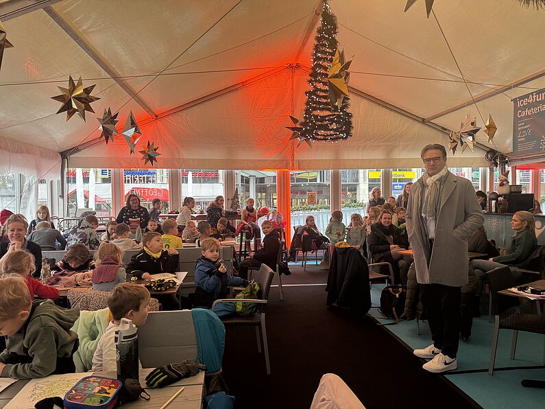 Im Bistro der der neu.sw Eislaufhalle sitzen Schülerinnen und Schüler und hören die Begrüßung von neu.sw Geschäftsführer Ingo Meyer.