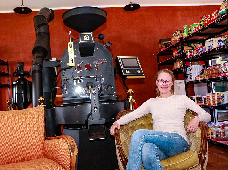 Antje Hornburg-Zeneli sitzt in einem gelben Sessel vor einer schwarzen Kaffeeröst-Maschine.