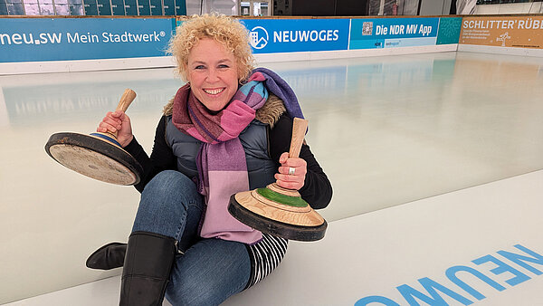 Moderatorin Friederike Witthuhn sitzt auf der Eisfläche der neu.sw Eislaufhalle und zeigt lächelnd zwei Eisstöcke in die Kamera.