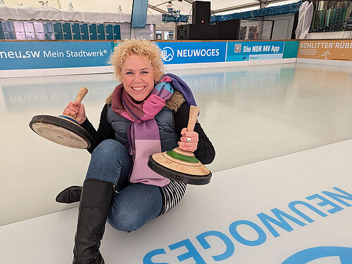 Moderatorin Friederike Witthuhn sitzt auf der Eisfläche und hält lächelnd zwei Eisstöcke in die Kamera.