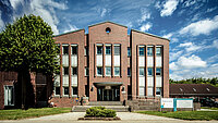 Frontansicht neu.sw Hauptgebäude. Blick auf Haupteingang. Rot-brauner Klinkerbau vor blauem Himmel mit weißen Wolken.