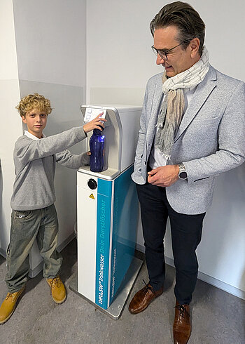 Schüler und neu.sw Geschäftsführer Ingo Meyer am weiß-türkis-farbenen neu.sw Wasserspender. Schüler füllt Wasser in eine blaue Trinkflasche.