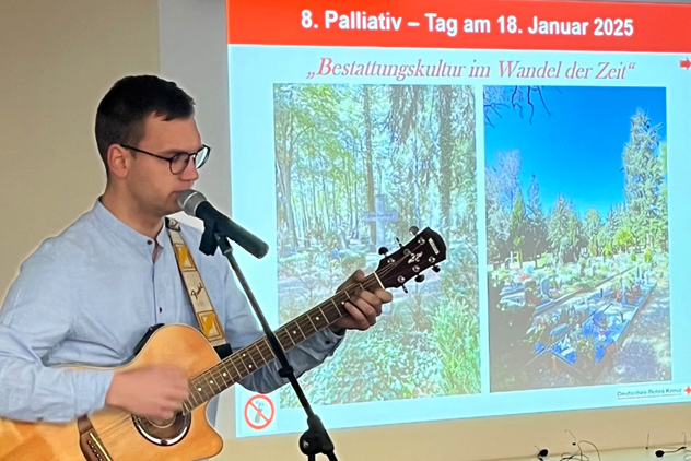 Sänger mit Gitarre vor einer Leinwand mit Tagungsbild. Es zeigt zwei Friehöfe und trägt die Überschrift "Bestattungskultur im Wandel der Zeit".